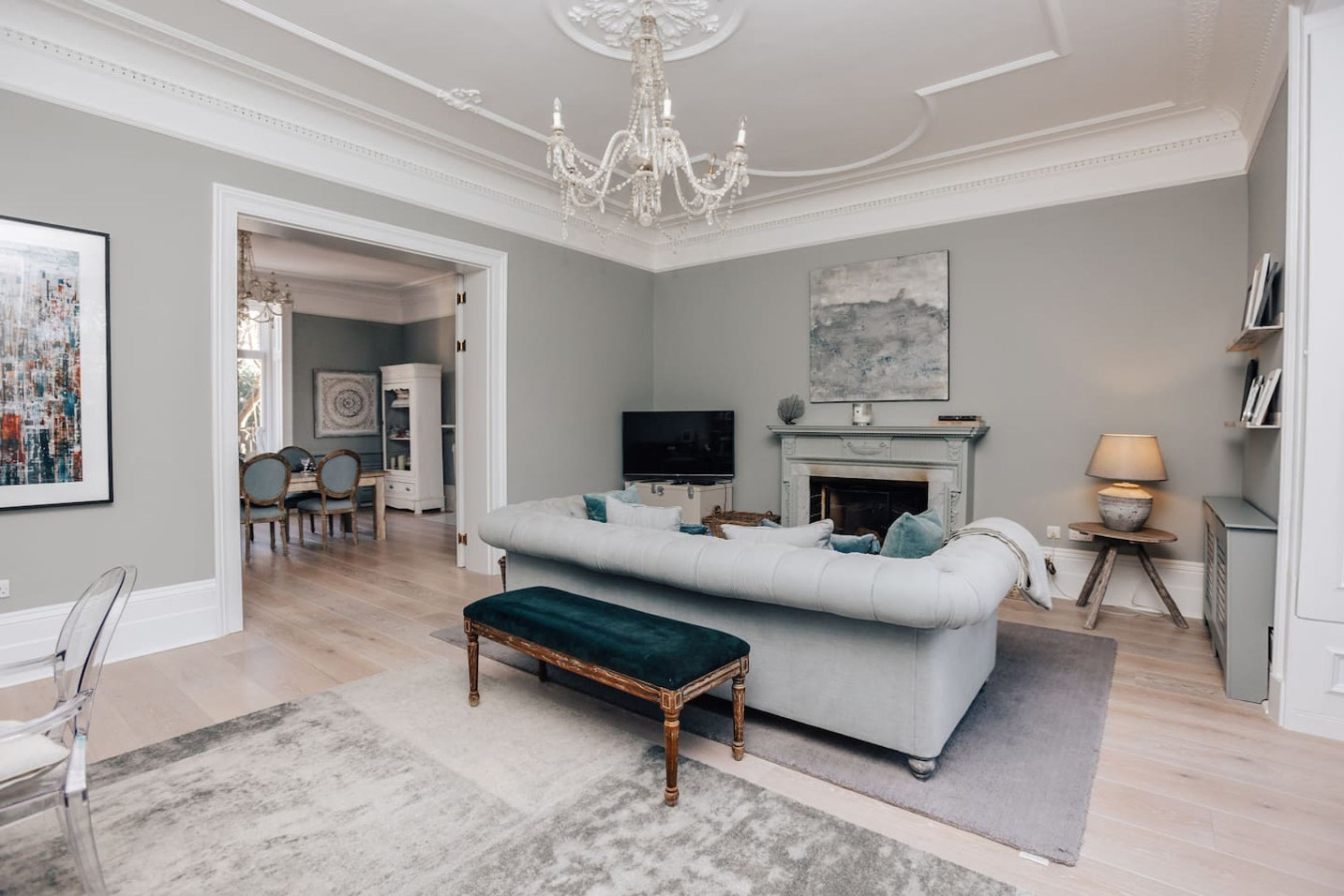 A spacious living room with a grey sofa, a wooden bench, a fireplace, and a chandelier.