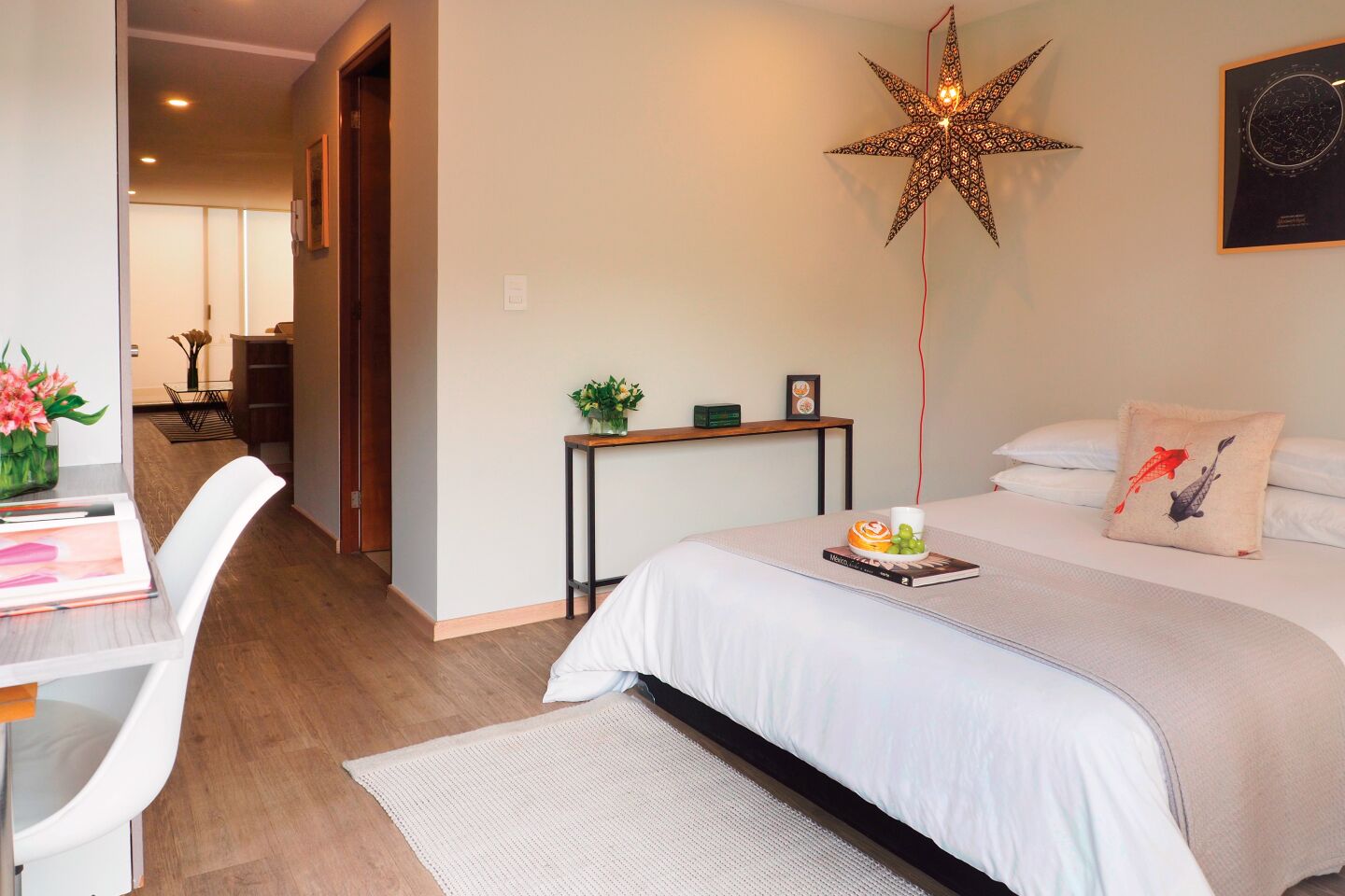 Quarto de hotel moderno com uma cama grande, uma escrivaninha com cadeira, uma luminária em formato de estrela e uma mesinha com uma planta e um porta-retrato.