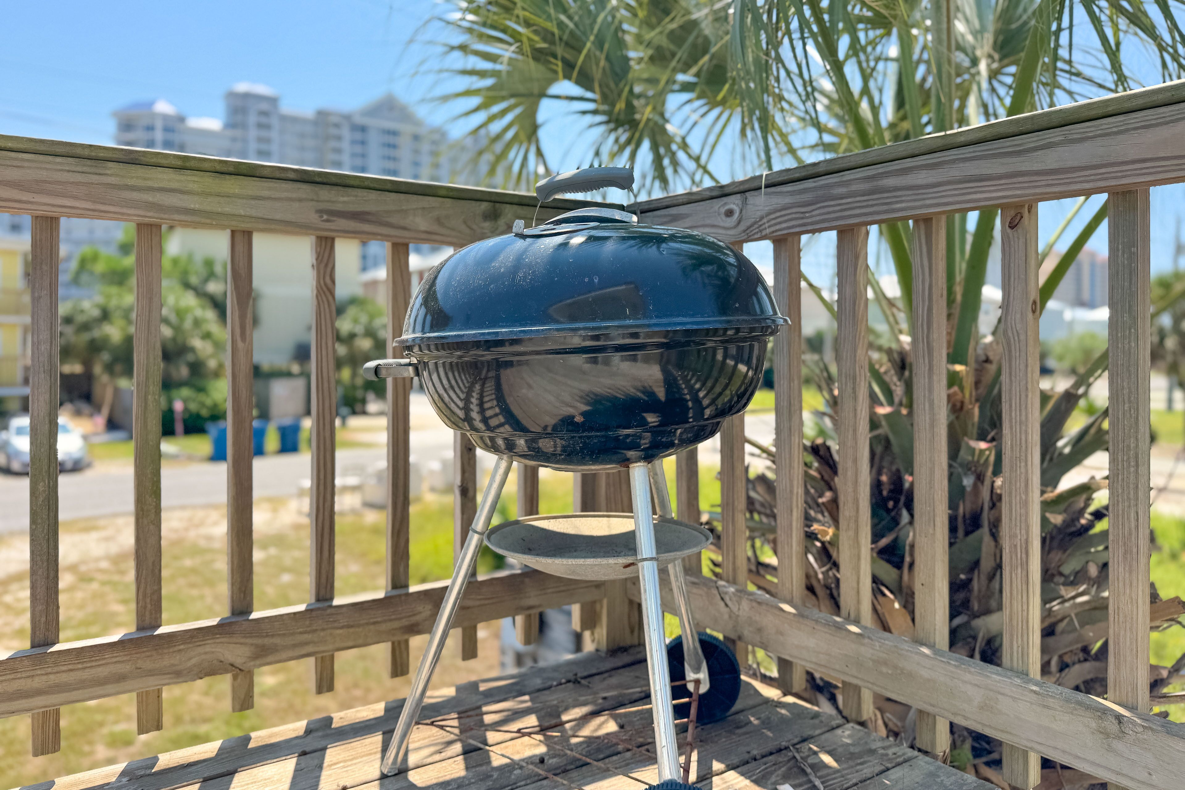 Una parrilla de barbacoa en una terraza de madera, con vista a palmeras y edificios al fondo.