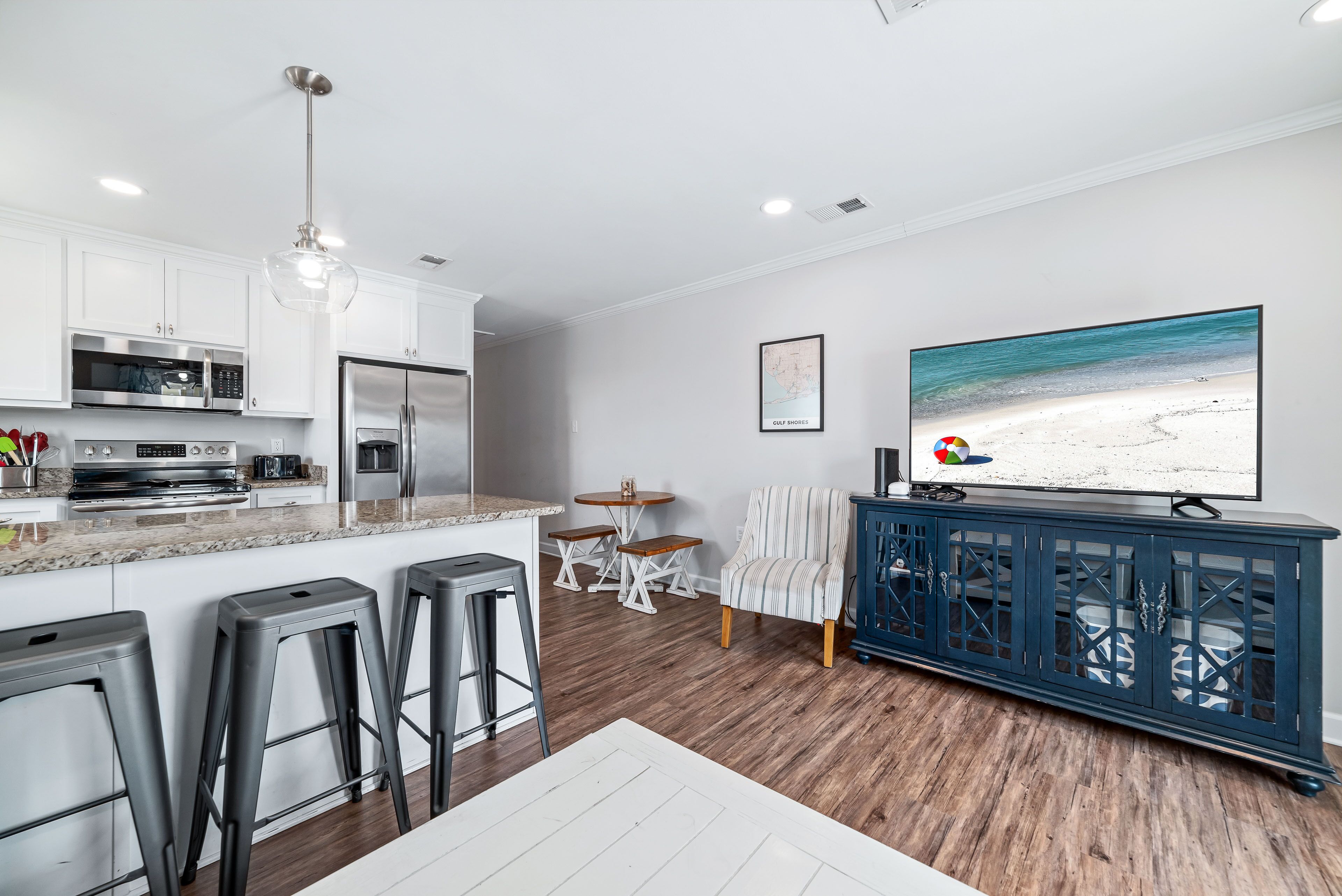 Een moderne keuken met roestvrijstalen apparatuur, een eethoek met een houten tafel en barstoelen, en een grote flatscreen-tv waarop een strandscène te zien is.