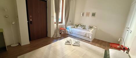 A bedroom with a bed, a nightstand, a chair, a window with curtains, a radiator, and a door.