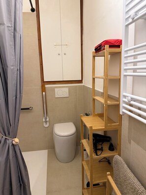 A small bathroom with a toilet, a wooden shelf unit, a towel rack, and a shower curtain. - Tuxedo Apartment your relaxation a stone's throw from downtown (Verbania)