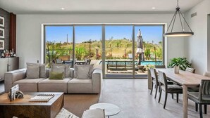 Ein modernes Wohnzimmer mit einem großen Schiebeglasfenster, das zu einem Poolbereich führt, einem Esstisch mit Stühlen und einem Sofa mit Kissen.