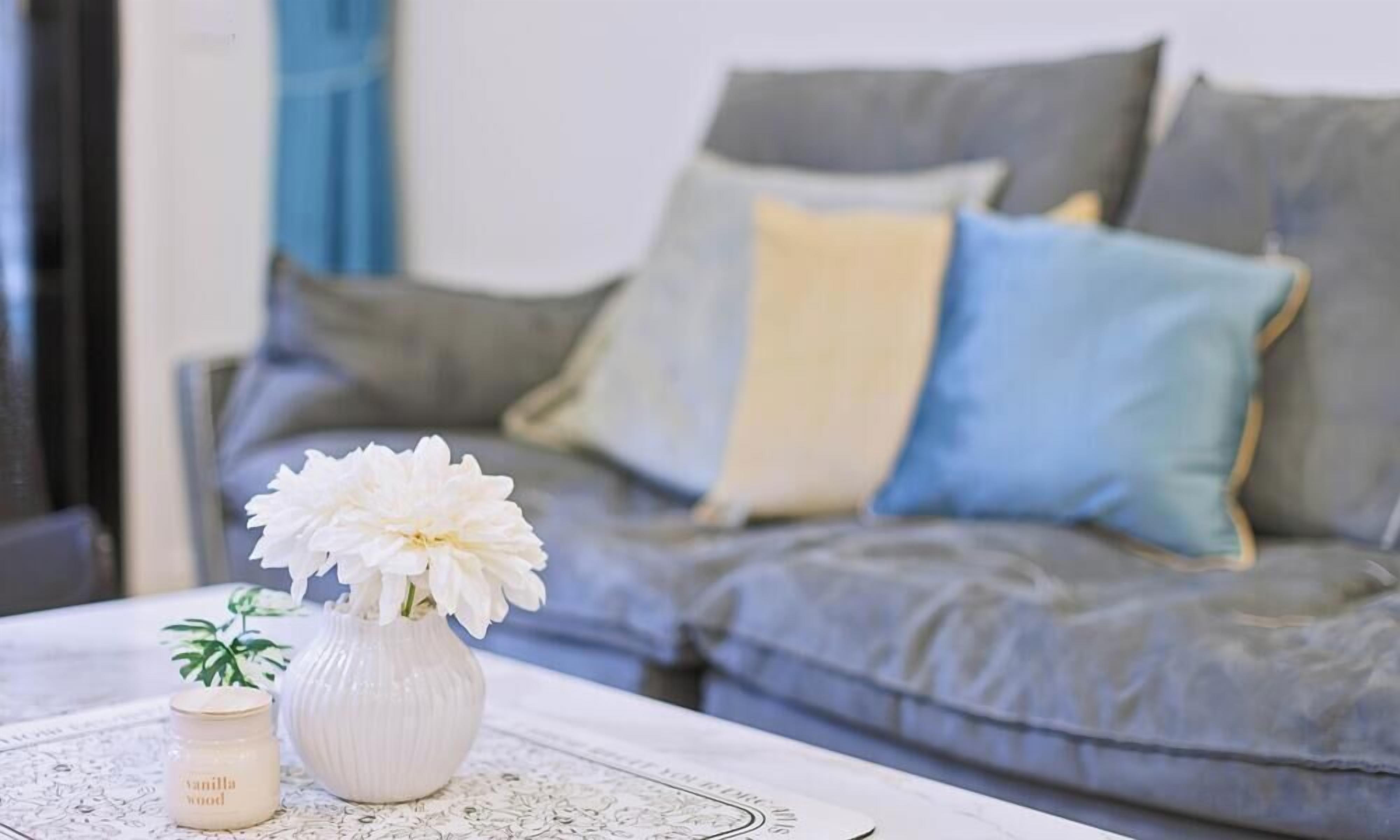 Ein Wohnzimmer mit grauem Sofa, einer weißen Vase mit Blumen und einer kleinen Kerze auf einem Tisch.