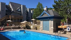 A swimming pool with lounge chairs and umbrellas, a small building with a blue door, and a staircase leading to a balcony.