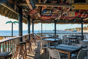 Un restaurante al aire libre con mesas y sillas de madera, techo de paja y vistas al mar.