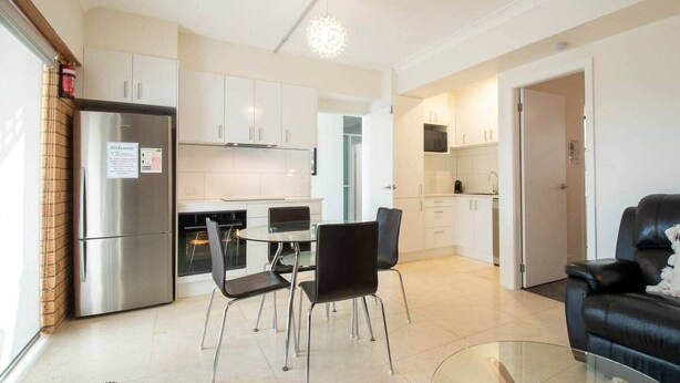 Een moderne keuken met roestvrijstalen apparatuur, een eethoek met een glazen tafel en zwarte stoelen, en een leren bank in een lichte woonkamer.