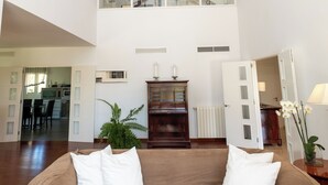 A spacious living area with a high ceiling, wooden beams, a sofa with white cushions, a plant, and a view of the kitchen and dining area. - Sa Roqueta – Beautiful villa with pool in the heart of Mallorca (Inca)
