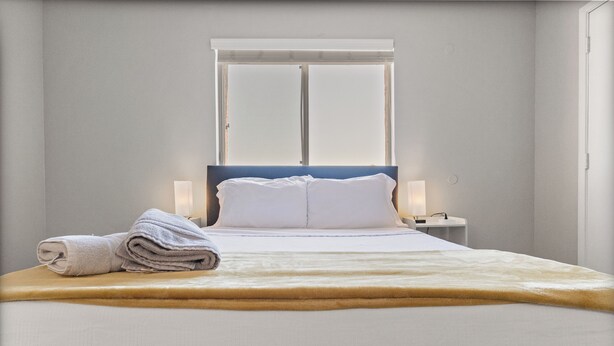 A neatly made bed with white linens and a folded towel on the side, a bedside table with a lamp, and a window with blinds. - Pool -Gym- Great for Mid/Long Stays (Mesa)
