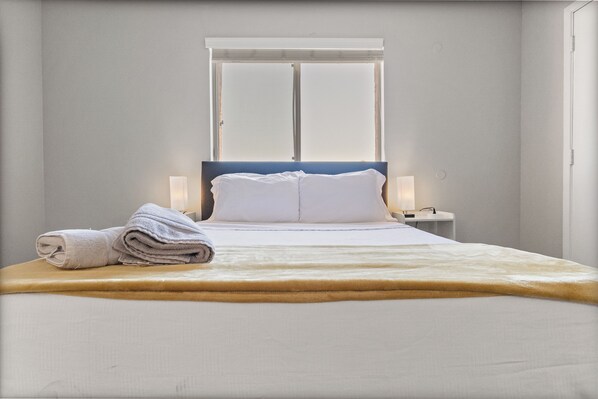 Un lit bien fait, avec du linge de lit blanc et une serviette pliée posée sur le côté, une table de chevet avec une lampe et une fenêtre avec des stores.