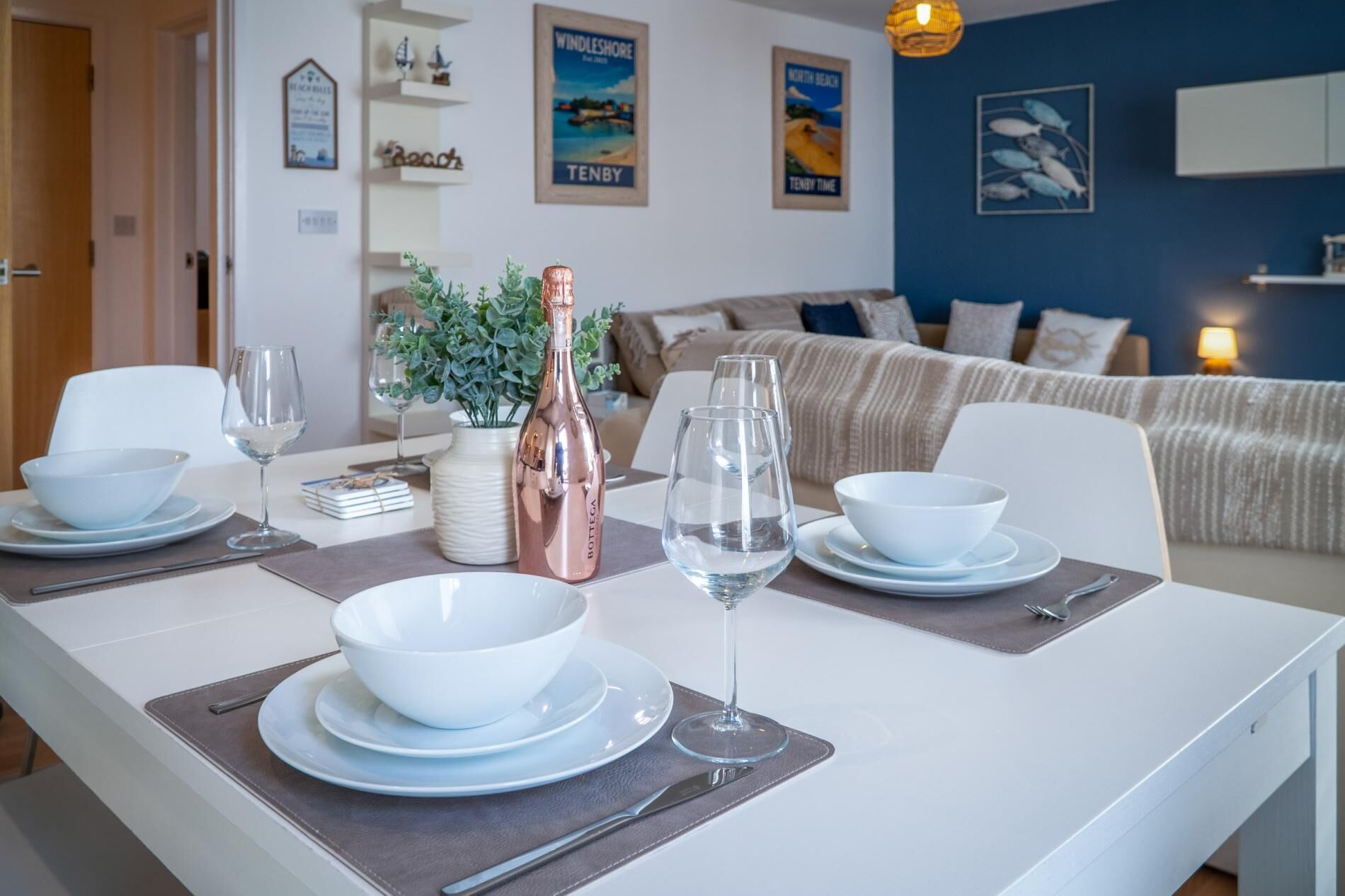 A dining area with a white table set for two, featuring a bottle of wine, two wine glasses, and white bowls and plates.