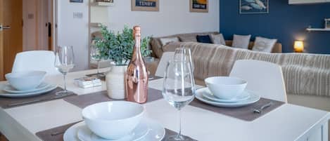 A dining area with a white table set for two, featuring a bottle of wine, two wine glasses, and white bowls and plates.
