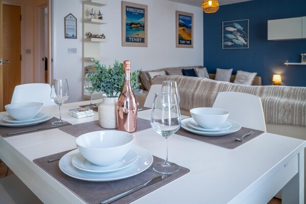 A dining area with a white table set for two, featuring a bottle of wine, two wine glasses, and white bowls and plates.