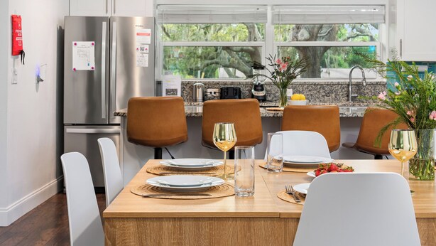 Une cuisine moderne avec des meubles blancs, des appareils en acier inoxydable et un coin repas composé de chaises marron et d’une table en bois.