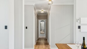 A hallway with a wooden floor, white walls, and a mirror on the far wall. There is a wooden table with a wine bottle and glasses on it.