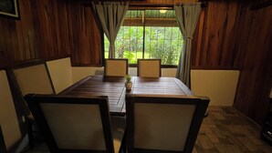 A wooden dining area with a table and chairs, a window with curtains, and a view of greenery outside. - Peaceful Jungle Home. River Access + Arenal Views (El Castillo)