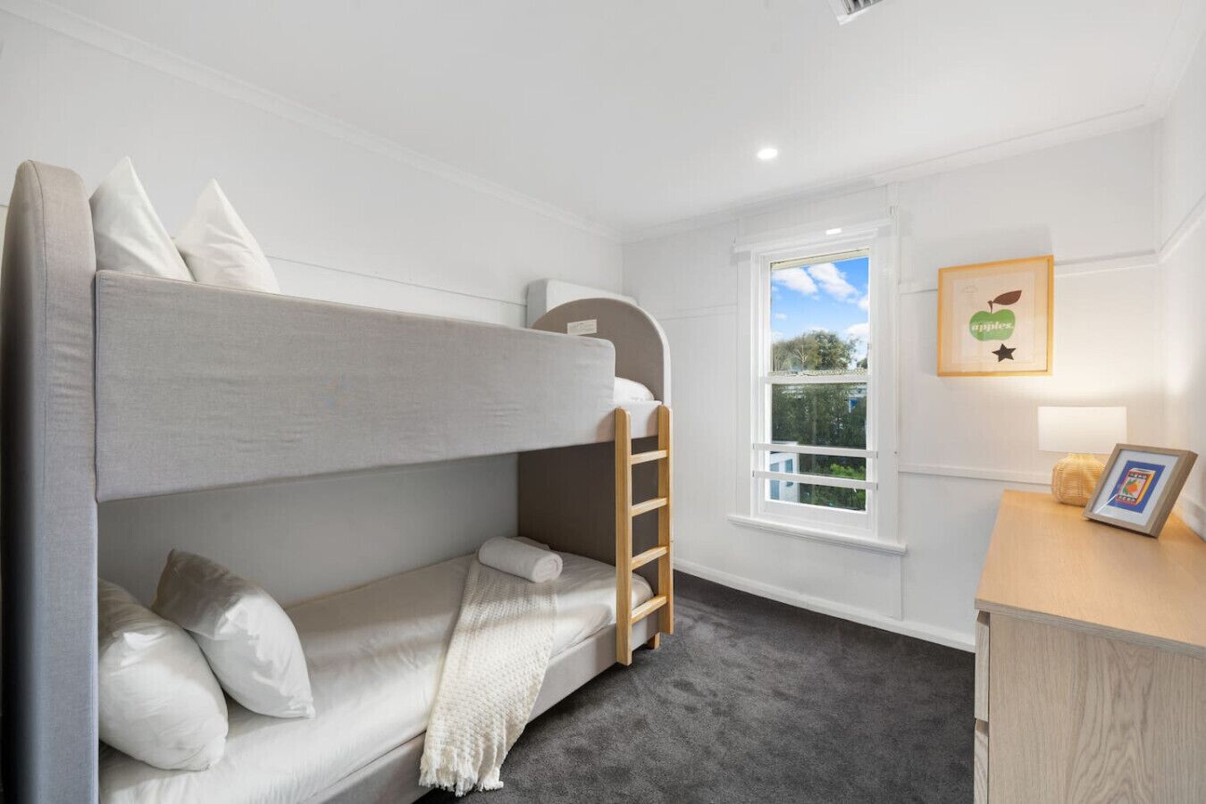 A bunk bed with pillows and a blanket, a wooden ladder, a window with a view, and a framed picture on a bedside table.