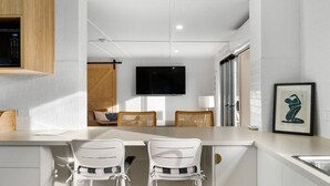 Cocina moderna con armarios blancos, microondas integrado y un televisor de pantalla plana grande montado en la pared. Hay dos taburetes blancos con detalles negros y una mesa de comedor con una silla de mimbre.