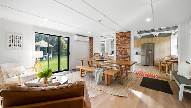 Una sala de estar moderna con un sofá de cuero marrón, un comedor con una mesa de madera y bancos, y una cocina con pared de ladrillo y electrodomésticos de acero inoxidable.