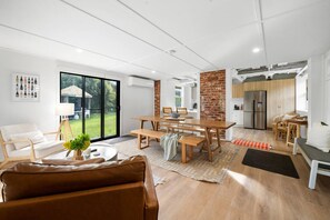 Ein modernes Wohnzimmer mit einem braunen Ledersofa, einem Essbereich mit einem Holztisch und Bänken sowie einer Küche mit Backsteinwand und Edelstahlgeräten.