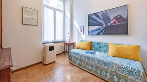 Una habitación con piso de madera, una cama con ropa de cama estampada, un radiador, una ventana con cortinas y un cuadro de un puente.