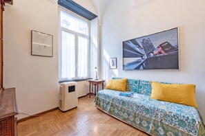 Una habitación con piso de madera, una cama con ropa de cama estampada, un radiador, una ventana con cortinas y un cuadro de un puente.