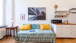 Un dormitorio con cama, mesita de noche, lámpara, marco de foto, estante con frascos, cafetera y ventana.