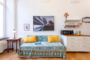 Un dormitorio con cama, mesita de noche, lámpara, marco de foto, estante con frascos, cafetera y ventana.