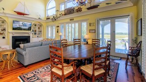 A spacious living room with a fireplace, a large dining table surrounded by wooden chairs, a sofa, and a television mounted on the wall.