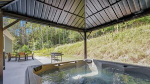 A hot tub with a covered patio, outdoor furniture, and a view of a forested hillside.