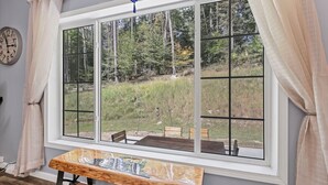 A room with a large window offering a view of a forested area, a wooden bench, and a clock on the wall.