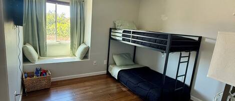 Une pièce avec un lit superposé, une fenêtre avec des rideaux, un panier en osier contenant divers objets et un téléviseur fixé au mur.