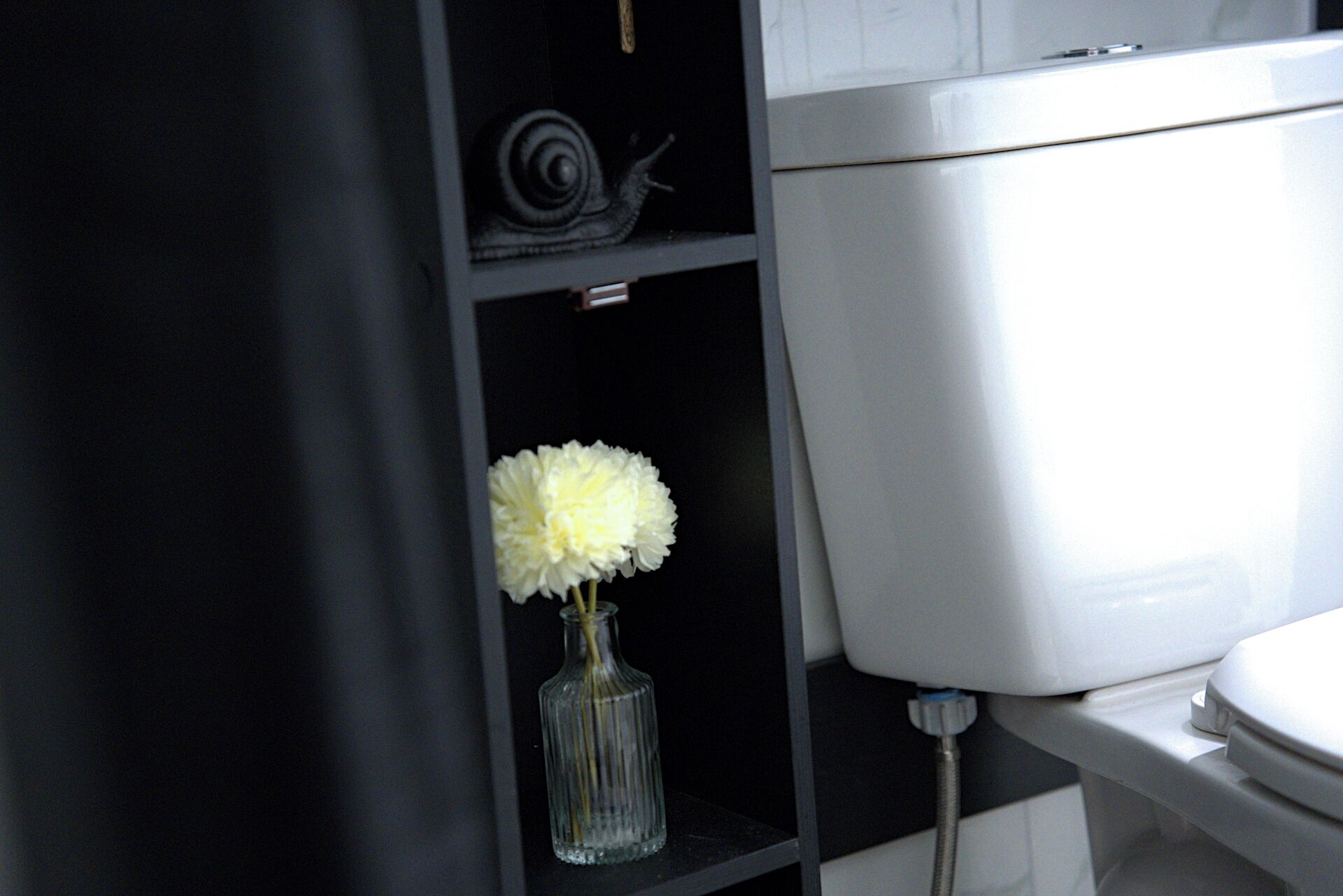 Casa de banho com sanita, um vaso de vidro transparente com flores brancas numa prateleira e um rolo de papel higiénico.