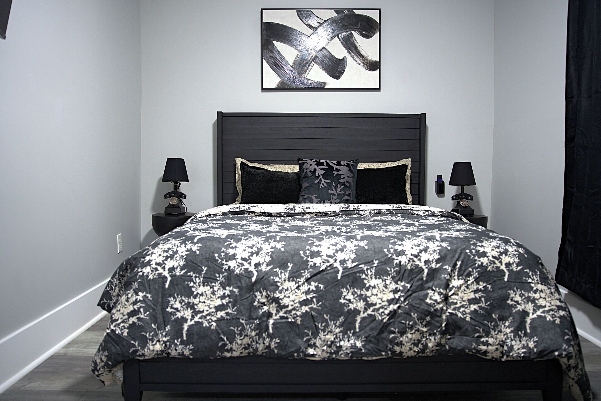 A bedroom with a black and white floral bedspread, a dark headboard, two bedside tables with lamps, and a framed abstract artwork on the wall.