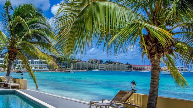 Blick auf ein Schwimmbecken, Palmen, das klare, blaue Meer und ein Hotel im Hintergrund.
