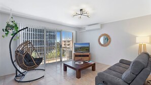Un salon comprenant un canapé, une table basse en bois, un téléviseur posé sur un meuble en bois, un fauteuil suspendu et un balcon offrant une vue sur les immeubles.