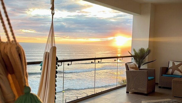 A balcony with a view of the sunset over the ocean, a hammock, and two wicker chairs with starfish cushions.
