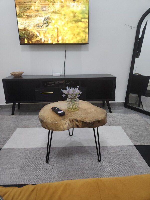 Un salon avec un téléviseur fixé au mur, une table basse en bois ornée d’un vase et d’une télécommande, un meuble de rangement noir pour appareils électroniques et un tapis à motifs.