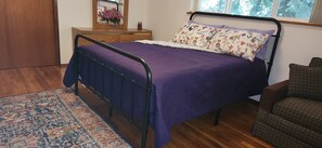 A bedroom with a metal bed frame, purple bedding, a floral pillow, a wooden dresser, a mirror, and a window with a view of trees.