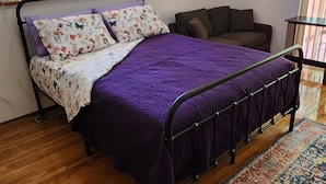 A bedroom with a bed, a sofa, a patterned rug, and a view of greenery through the window.