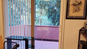 A room with a wooden floor, a black table with a chair, a small trash bin, and a door leading to an outdoor area with a red railing and greenery.