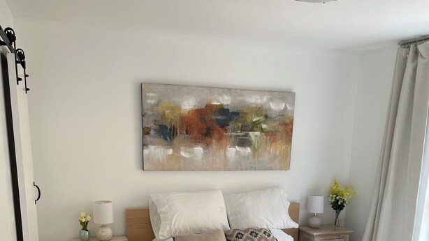 Une chambre à coucher avec un grand lit, une table de chevet, un tableau abstrait accroché au mur et une fenêtre avec des rideaux.