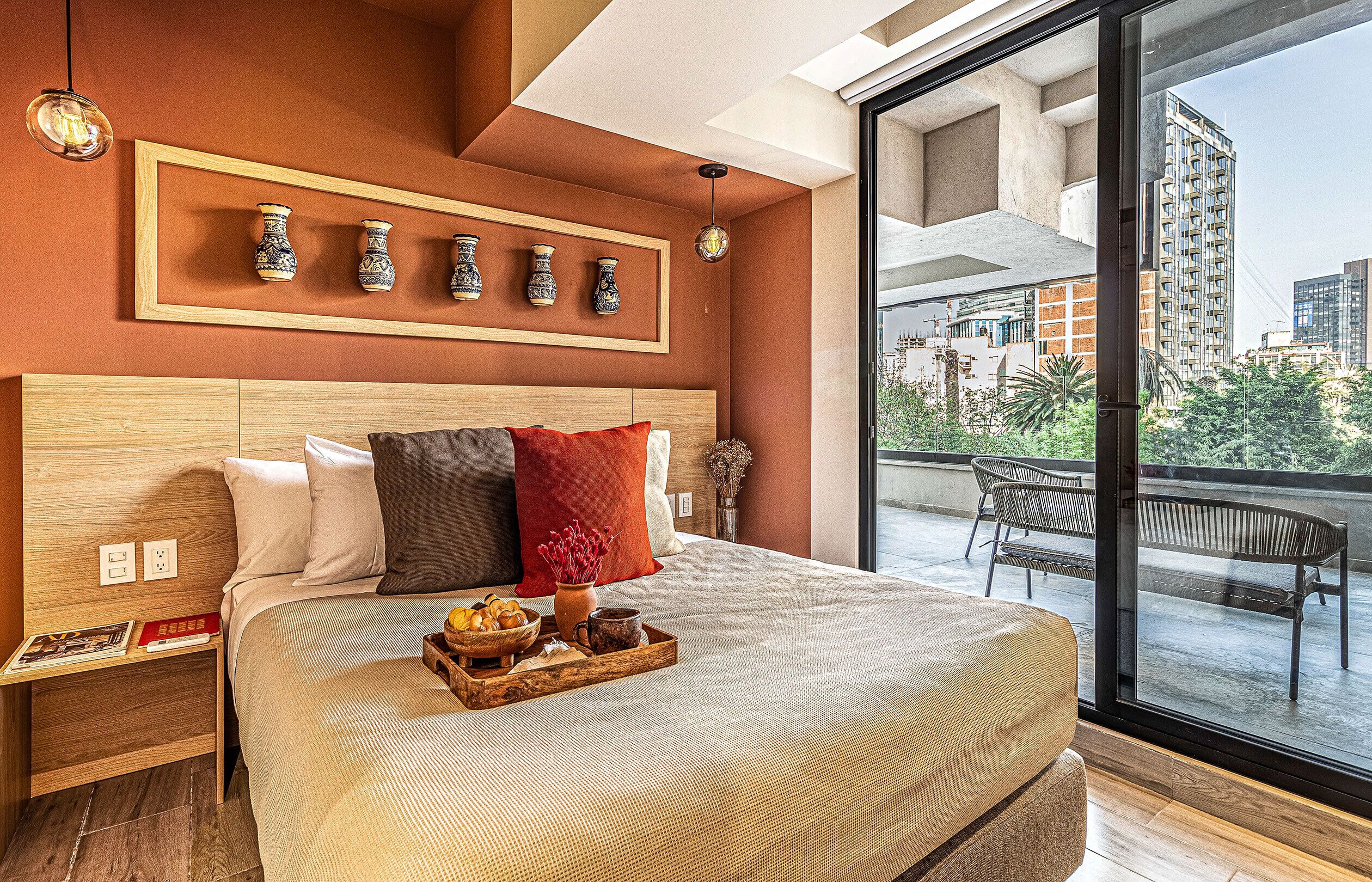Una habitación de hotel moderna con una cama grande, un cabecero de madera, una bandeja con alimentos para el desayuno y un balcón con vistas a la ciudad.