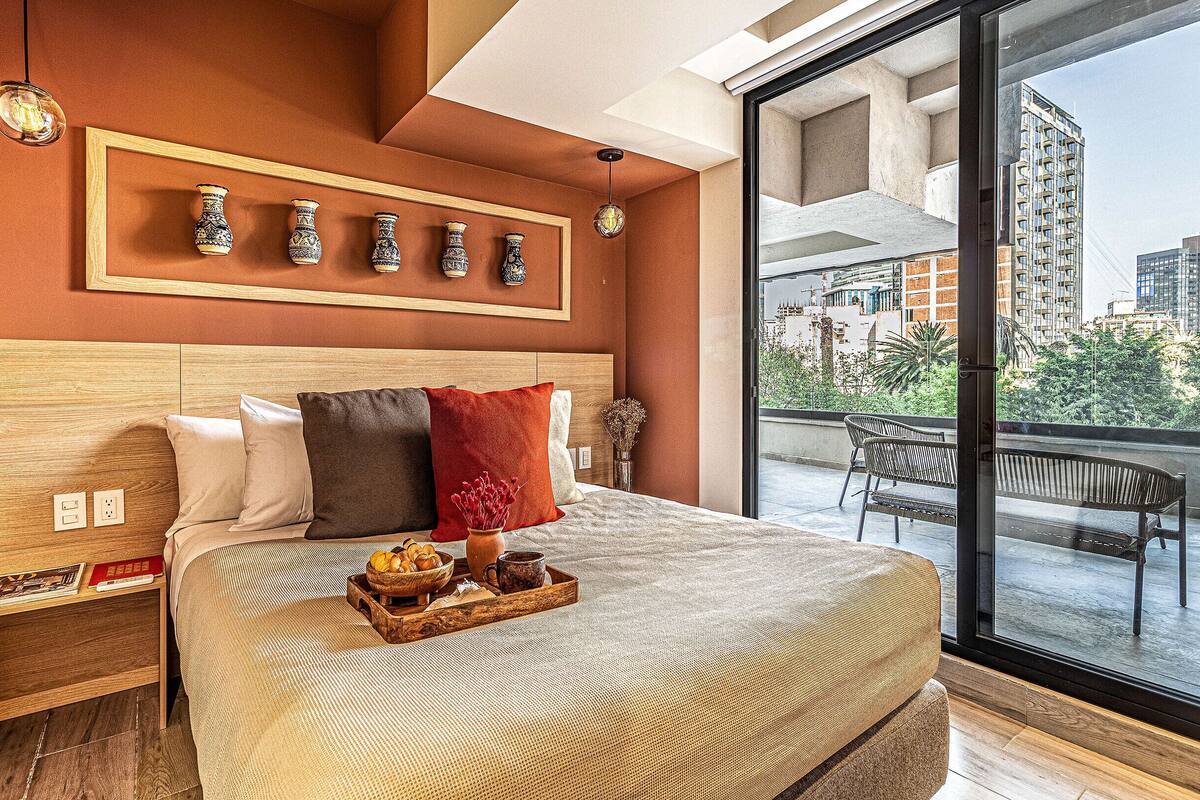 Une chambre d’hôtel moderne avec un grand lit, une tête de lit en bois, un plateau avec des articles pour le petit-déjeuner et un balcon offrant une vue sur la ville.
