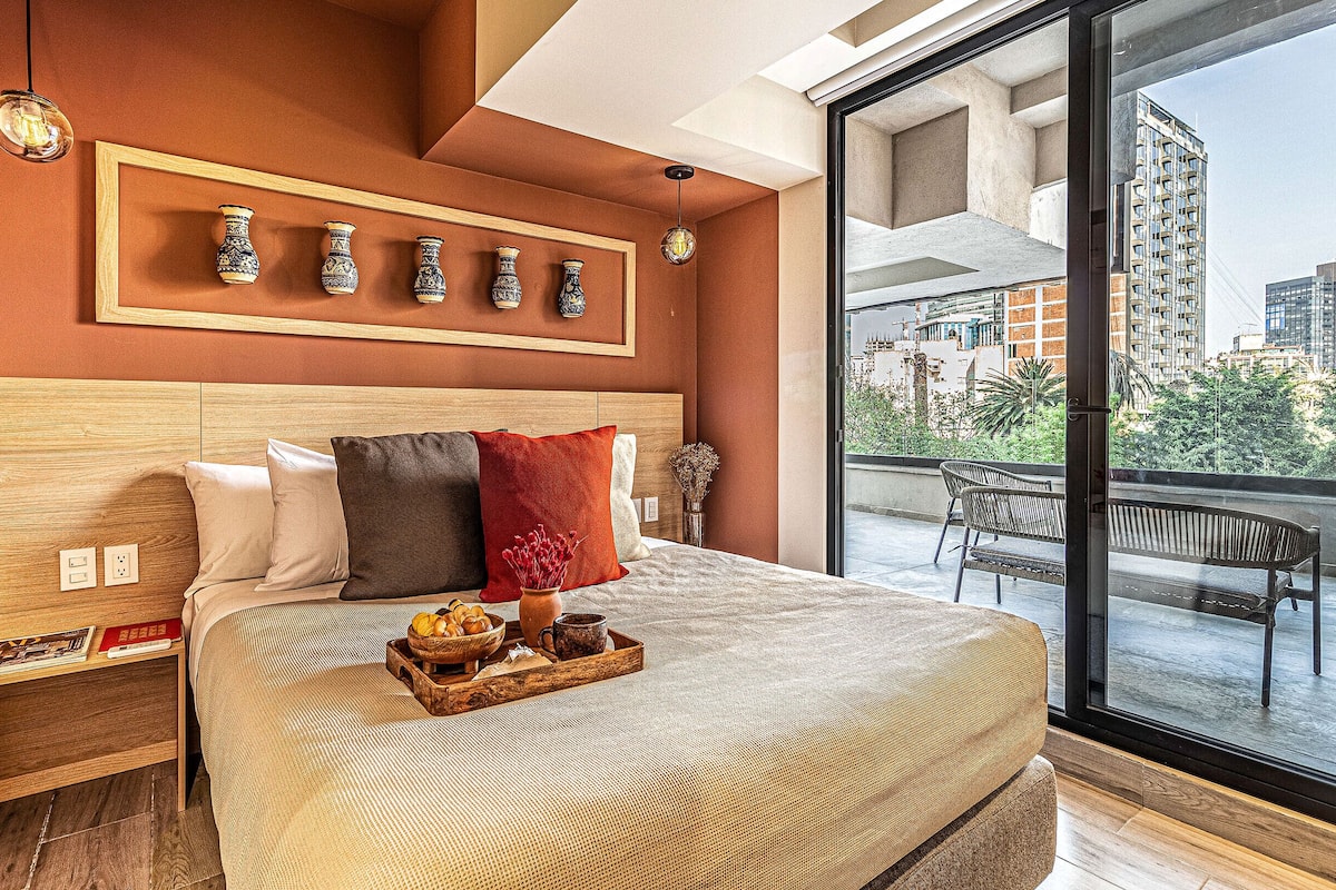 Una habitación de hotel moderna con una cama grande, un cabecero de madera, una bandeja con alimentos para el desayuno y un balcón con vistas a la ciudad.