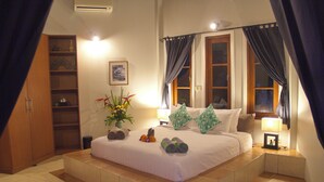 Habitación de hotel con una cama grande, una mesita de noche con lámpara, una estantería y una ventana con cortinas.