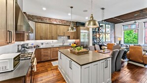 A modern kitchen with wooden cabinets, a large island, and a dining area with a view of greenery outside.