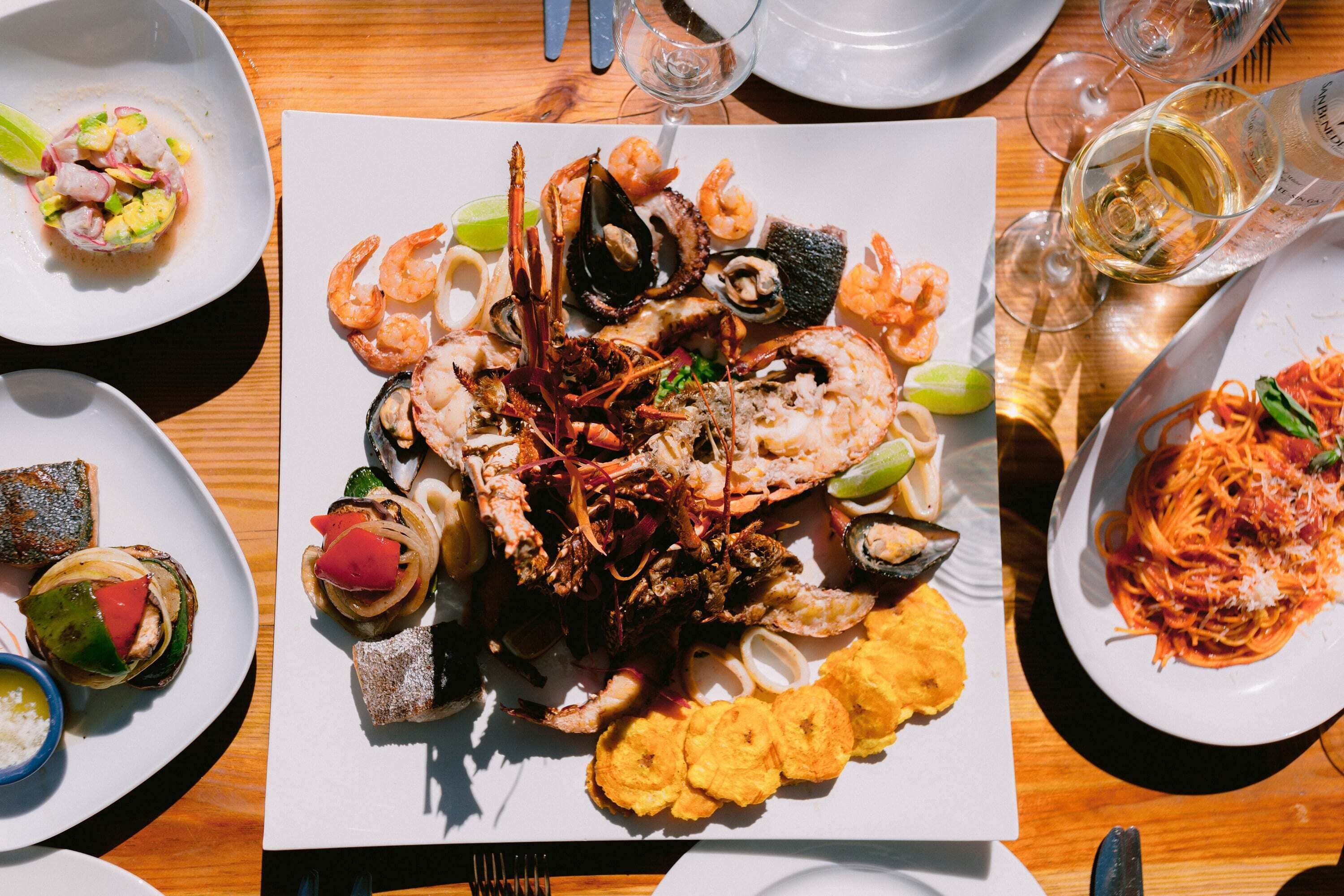 Un plateau de fruits de mer comprenant divers crustacés et légumes, accompagné d’un verre de vin blanc et d’une assiette de spaghettis.