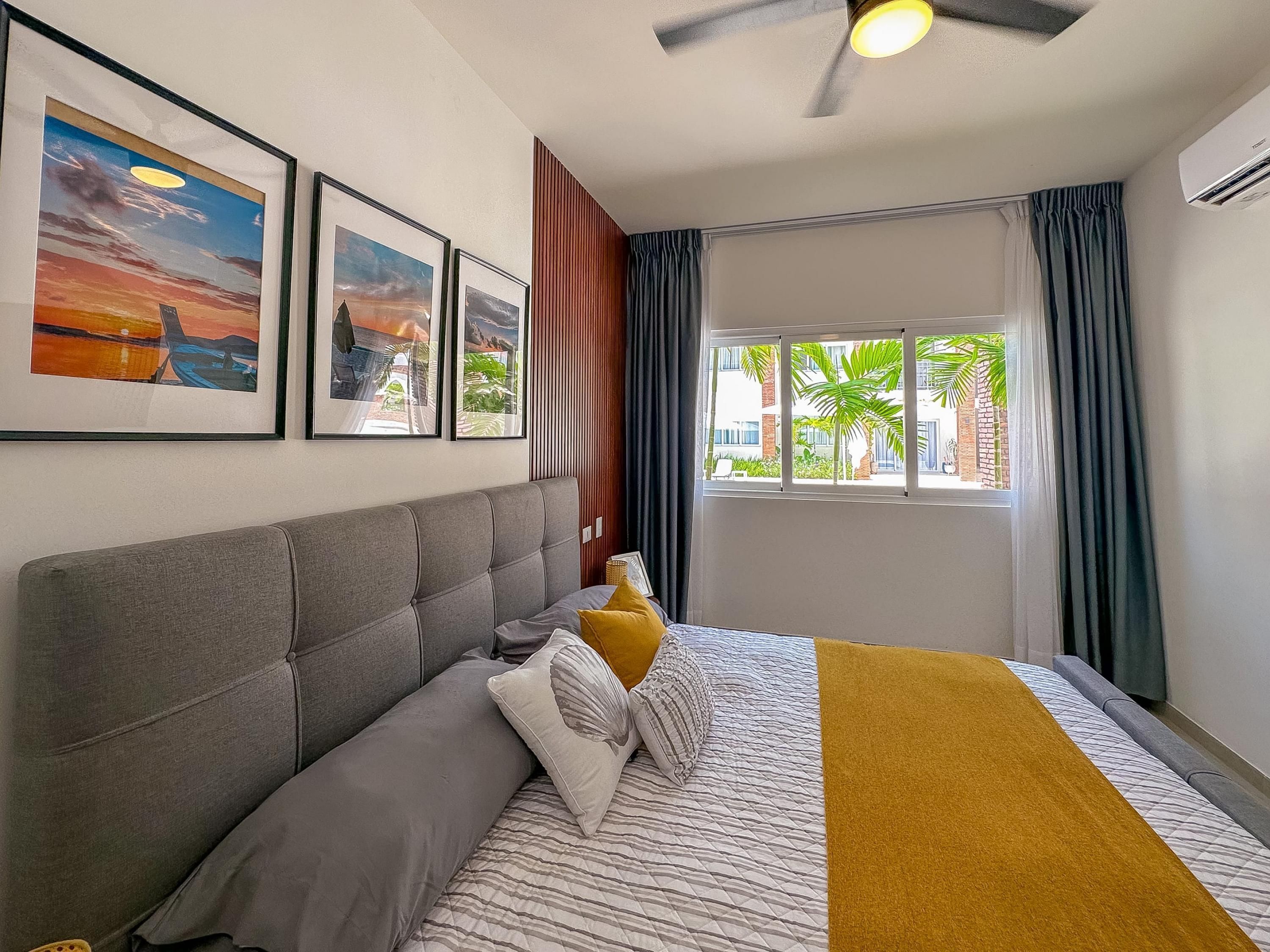 A neatly arranged bedroom with a bed, pillows, a headboard, a window with curtains, and framed pictures on the wall.