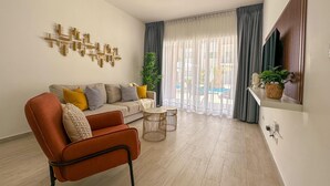 A modern living room with a sofa, armchair, and coffee table. There is a large window with curtains, a plant, and a view of the outdoors.